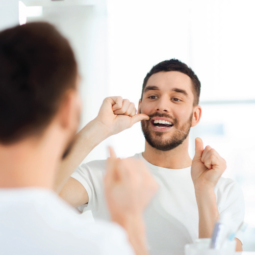 smiling young man with floss cleaning teeth and looking to mirror at home