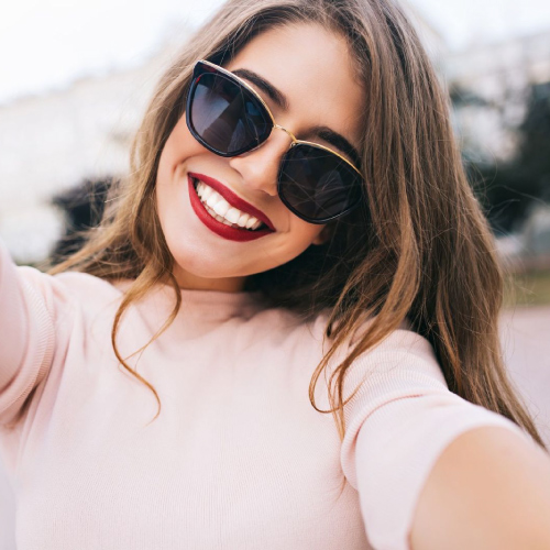 Closeup selfie-portrait of attractive girl in sunglasses with long hairstyle and snow-white smile in city.